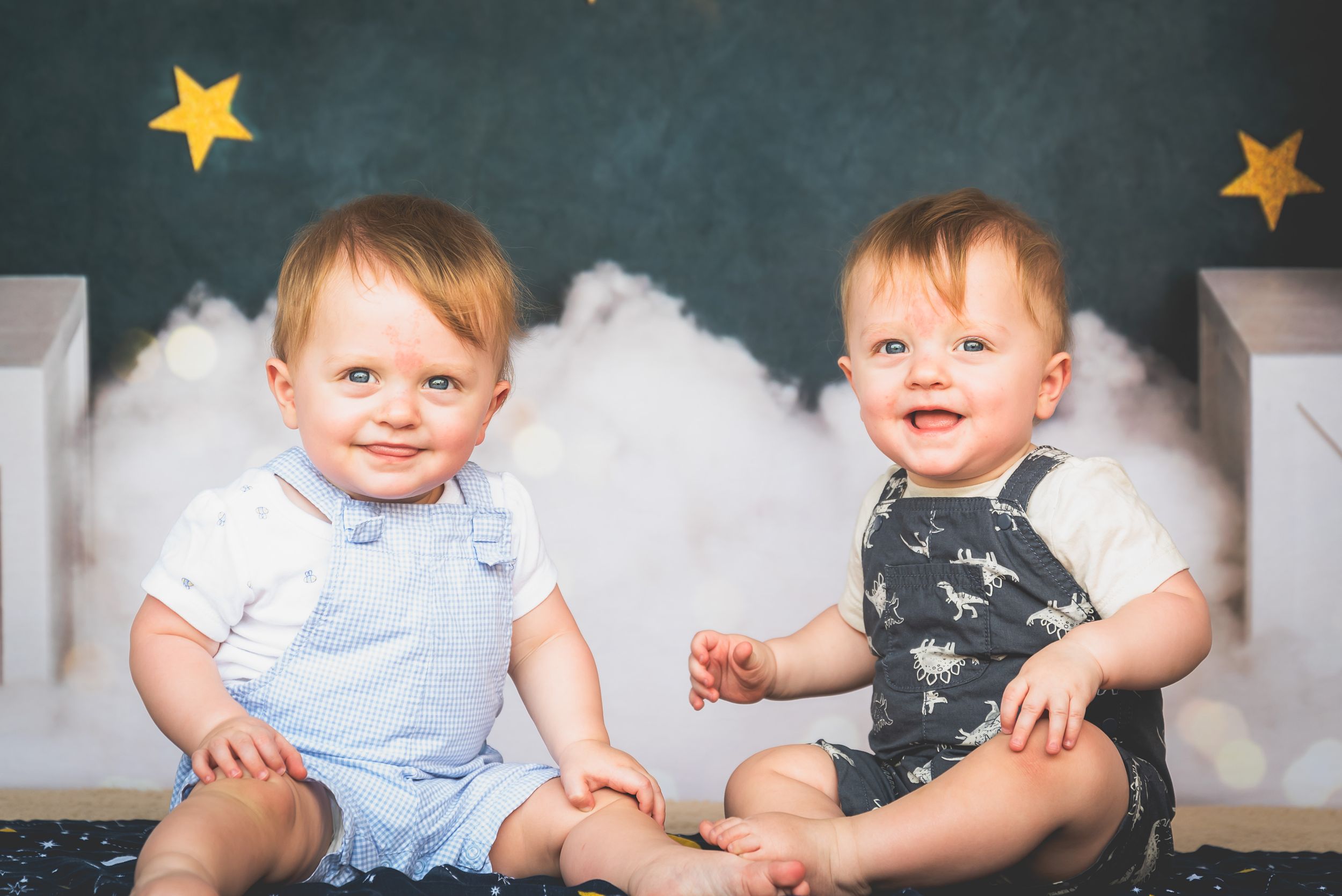 Two smiling baby twins sitting together in matching rompers against a starry night sky backdrop.