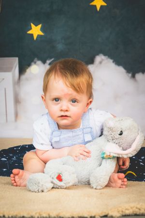 Adorable baby with blue eyes sits on a blanket hugging a gray stuffed elephant toy against a starry backdrop.