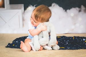 Adorable baby in diaper hugging a soft gray stuffed bunny toy while sitting on a star-patterned blanket.
