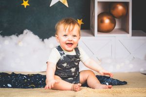 Smiling baby in denim overalls sits on a blanket with star and balloon decorations in the background.