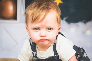 Adorable blonde toddler with blue eyes making a pouty face, wearing denim overalls, with a colorful backdrop.