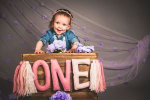 Smiling baby girl celebrating her first birthday beside a wooden crate with ONE sign, purple flowers, and pink tassels.