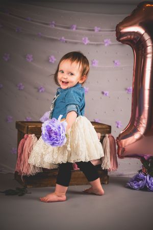Smiling baby girl in denim top and tutu skirt holds purple flower near rose gold number 1 balloon at first birthday photoshoot.