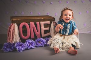 A joyful baby girl sits beside a wooden ONE sign with purple flowers for a first birthday photo shoot.
