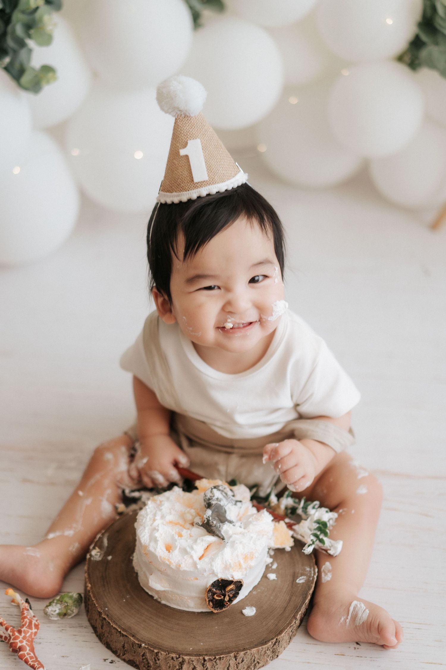 Baby in white outfit enjoys smashing and playing with birthday cake during cake smash photo session with white balloons.