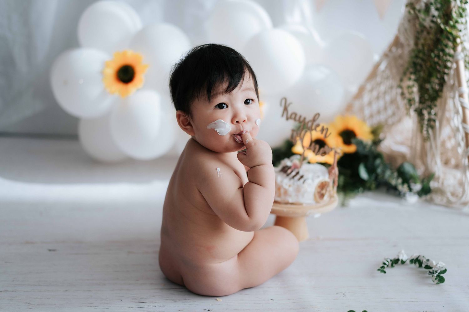A baby milestone photoshoot with sunflowers and white balloons in a rustic studio setting.