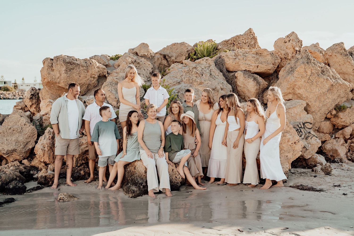 Wedding party members pose together on rocky beach cliffs at sunset in coordinated neutral-toned attire.