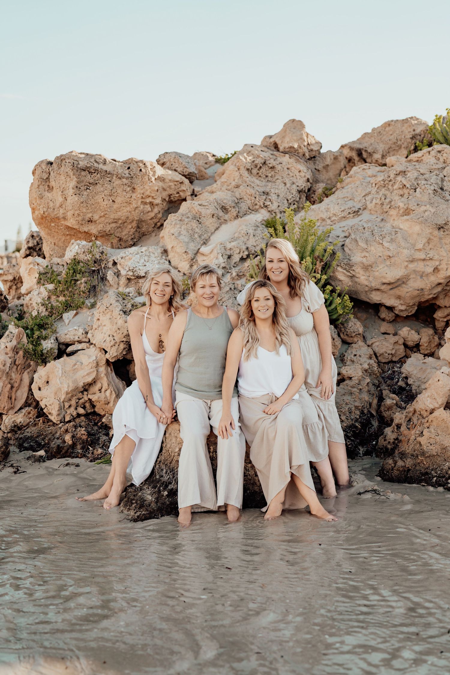 A group in coordinated neutral outfits sits together on a rocky beach with waves lapping at their feet.