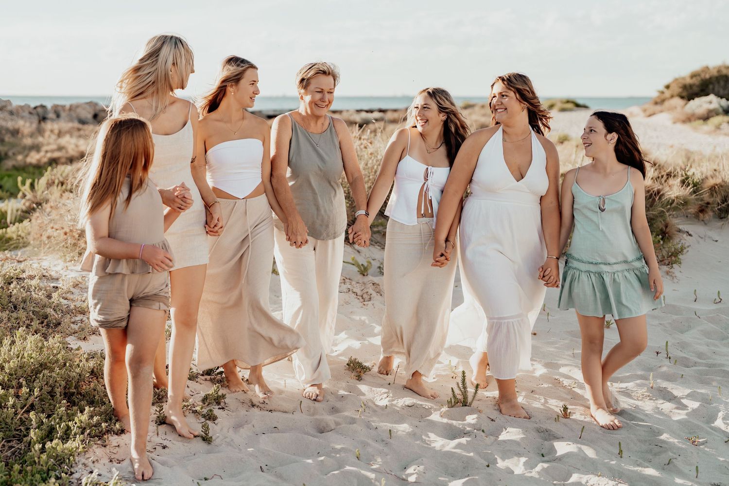 A group of people in light colored dresses and outfits walking together on a sandy beach at sunset.