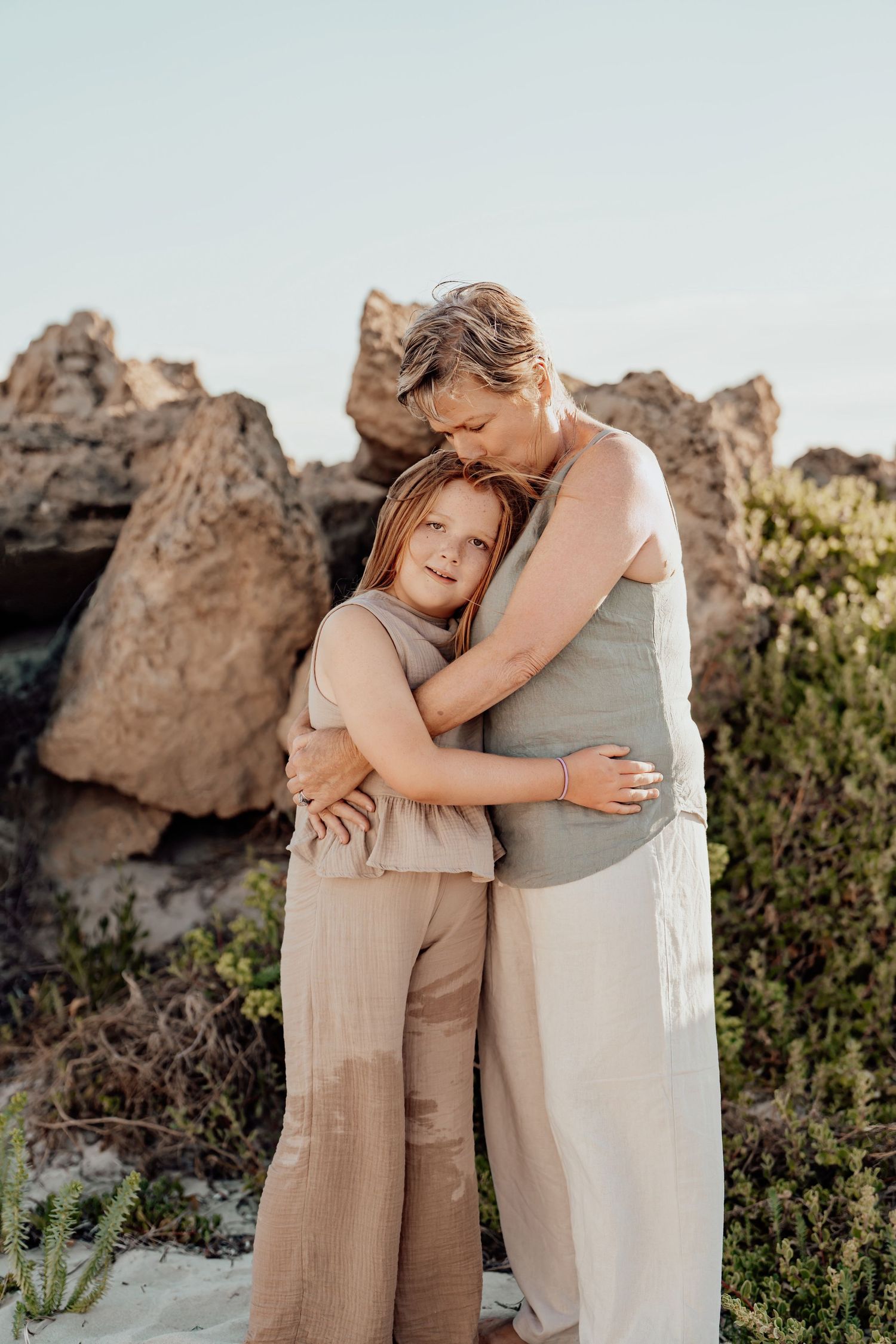 Two people in light beige outfits embrace tenderly against a backdrop of desert rocks during a golden hour photoshoot.