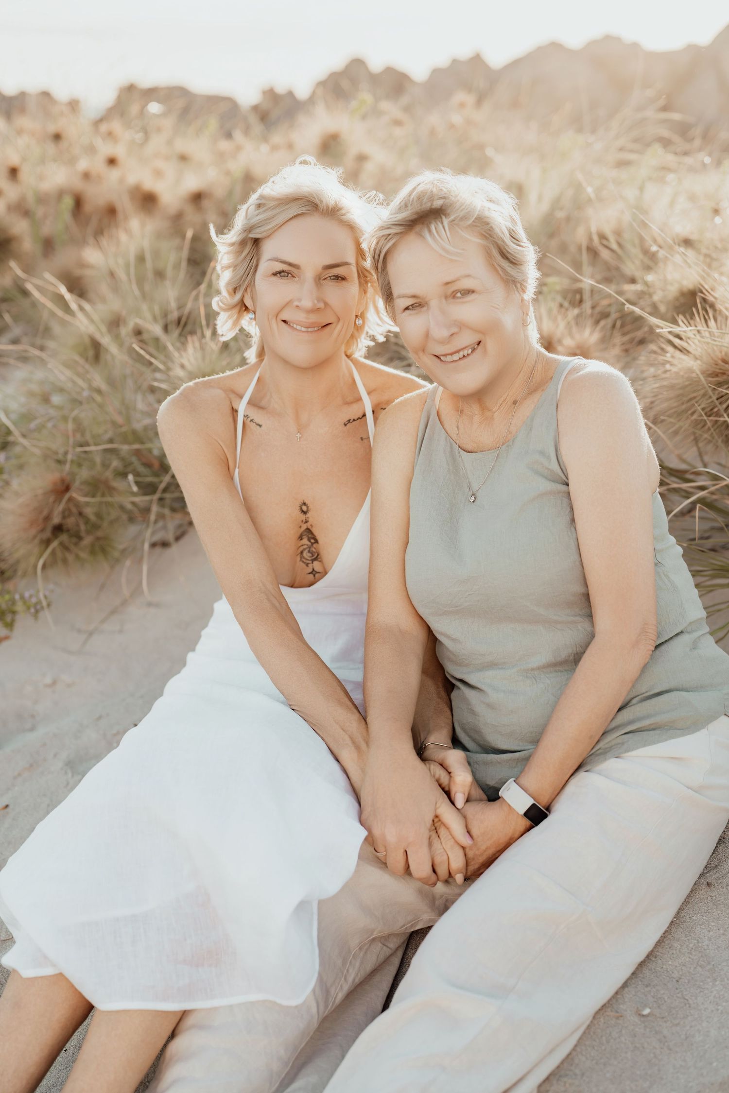 Two people share a tender moment together while sitting on sandy beach dunes in soft golden light.
