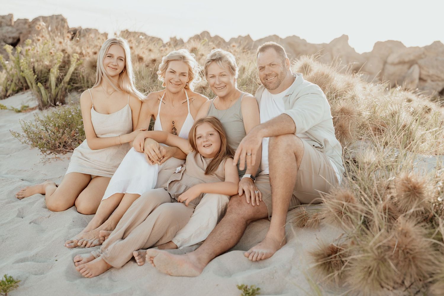 A group of people wearing coordinated neutral outfits sit together on a sandy beach at sunset.