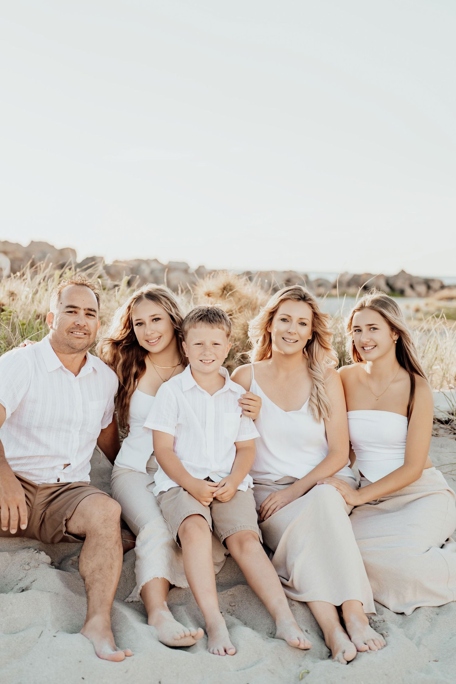 A family wearing coordinated white outfits sits together on a sandy beach during sunset.