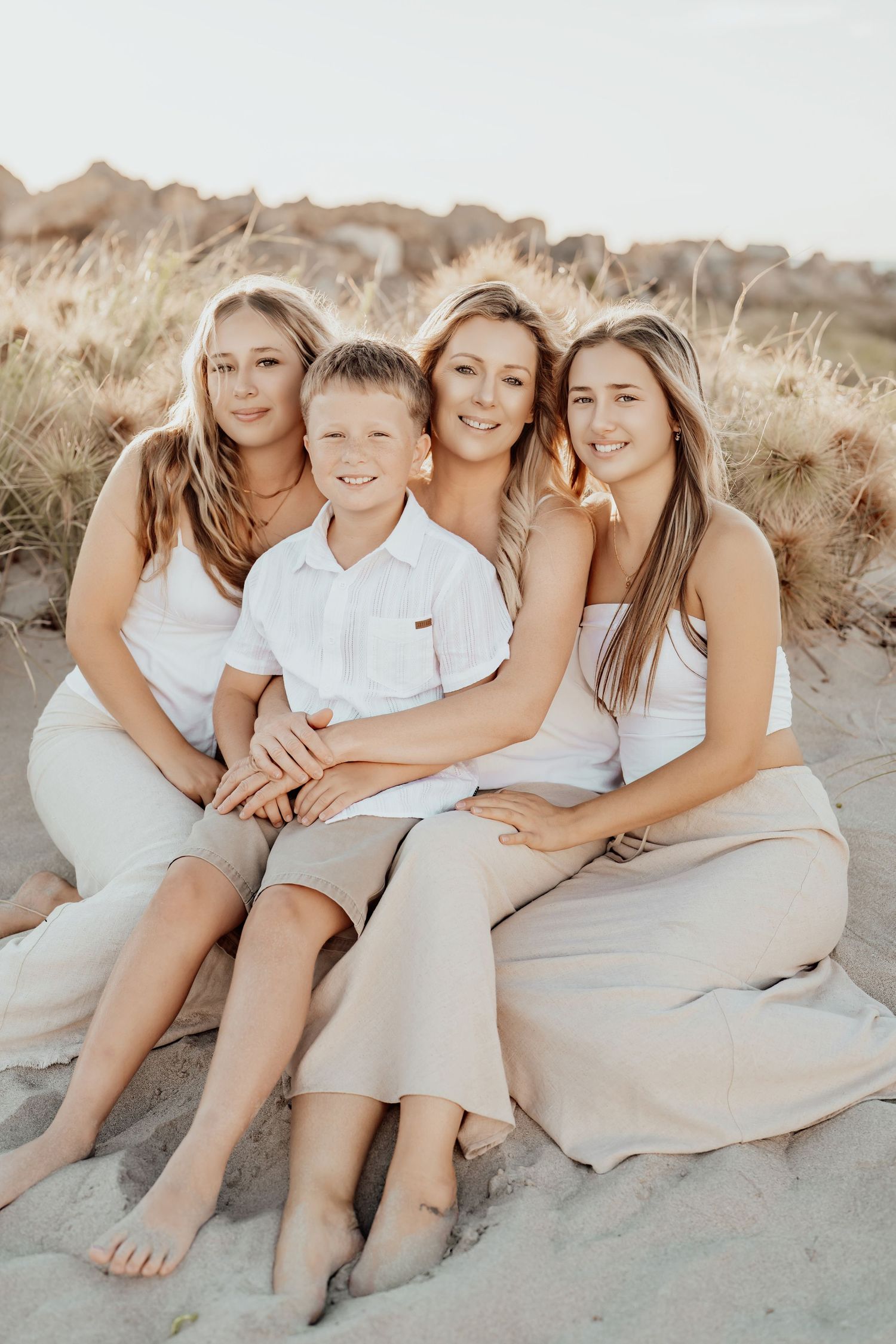 A family wearing white and beige sits together on a sandy beach during a warm sunset photoshoot.