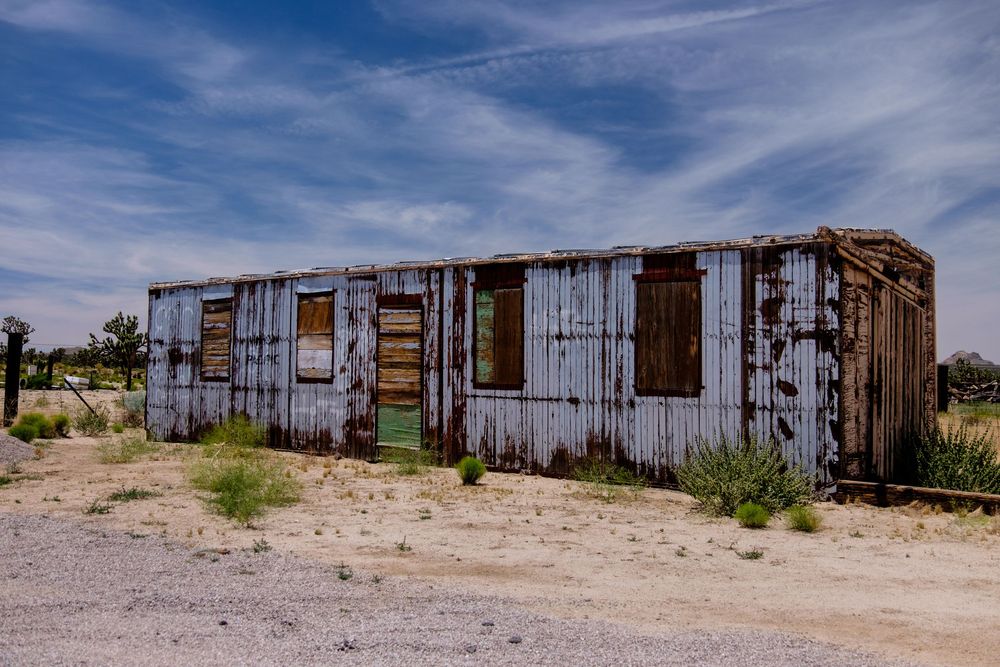 A series of abandoned rustic train cars sit in a desert landscape under blue skies with wispy clouds.