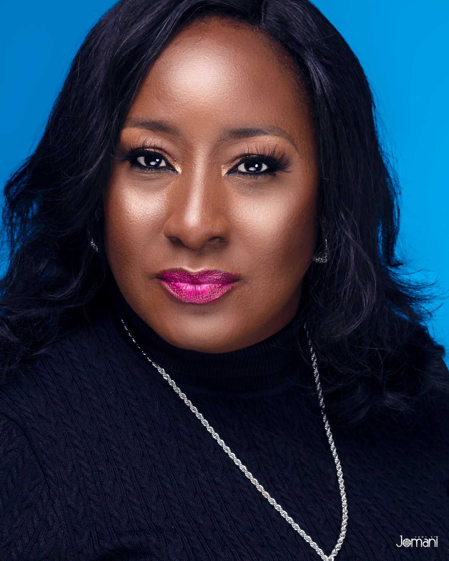 Professional headshot of a woman in black turtleneck with silver necklace against a blue background.