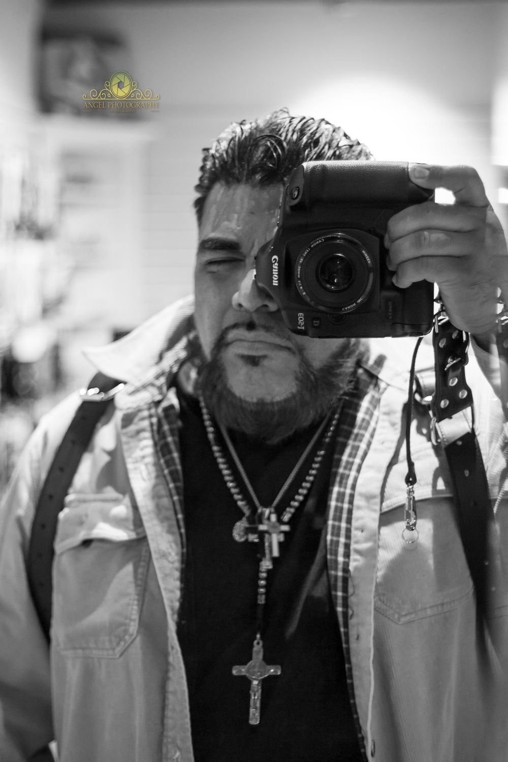 Black and white photo of a bearded man holding a Canon camera, wearing cross necklaces and a plaid jacket.