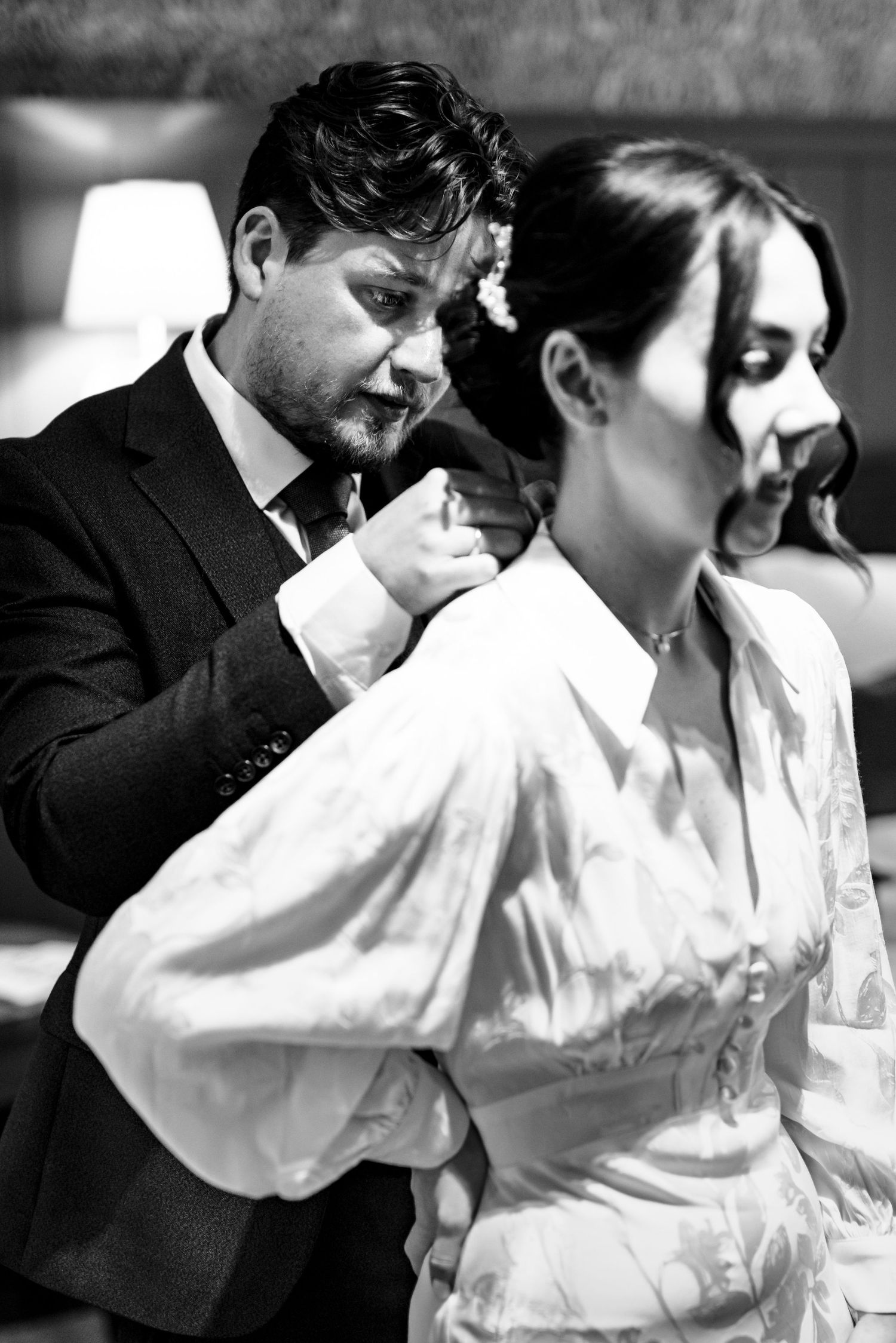A dramatic black and white photo shows a couple in formal attire, with him adjusting her dress from behind.