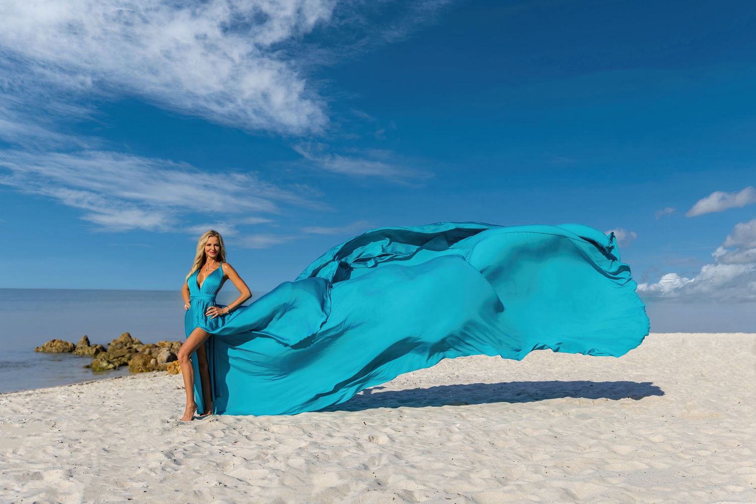 Flowing turquoise fabric billows dramatically in the wind on a pristine white sand beach against a bright blue sky.