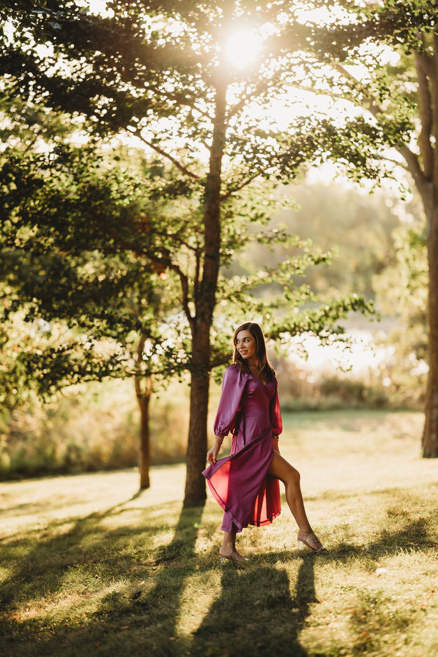 A person in a purple dress poses gracefully in a sunlit park with golden light streaming through the trees.