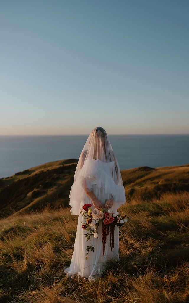 A bride in ruffled veil holds a bouquet purse, on a coastal cliff at sunset. Howdy Pictures - film wedding & elopement photographers in NZ