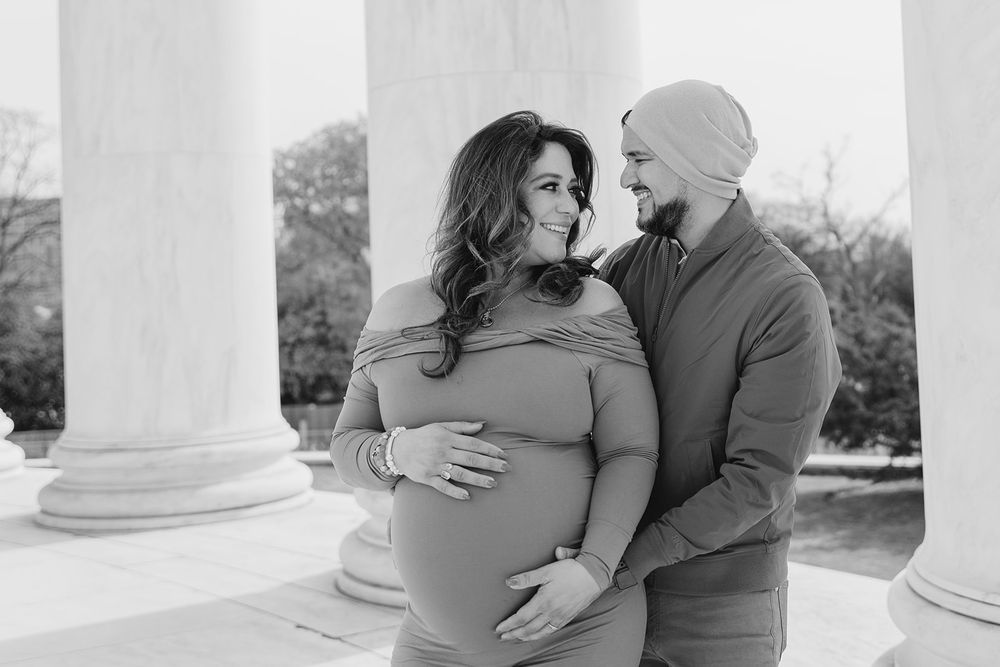 An intimate black and white portrait of an expecting couple sharing a tender moment between classical columns.