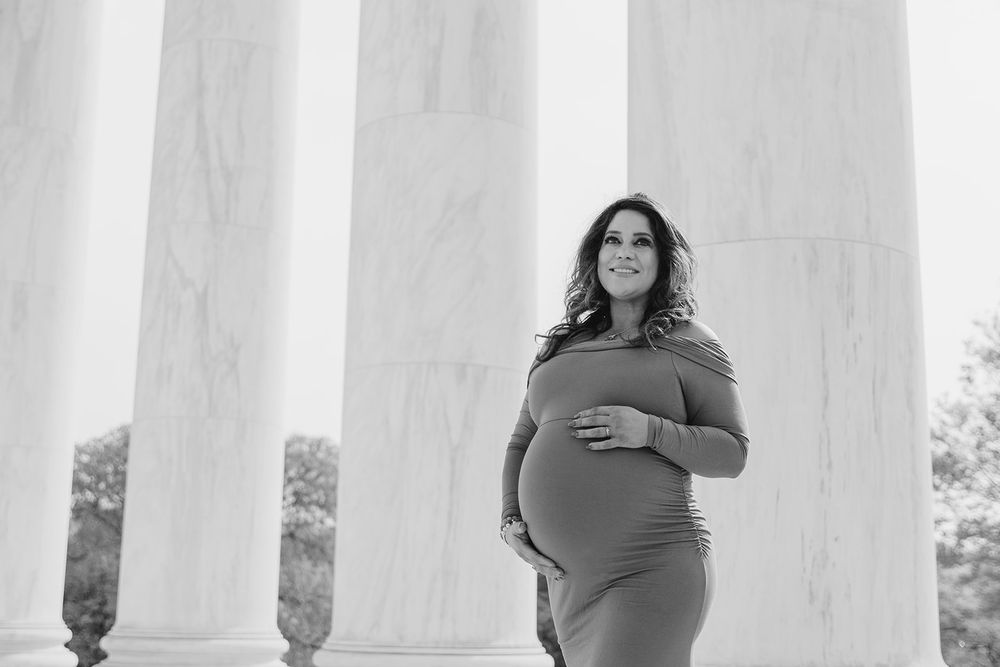 Black and white maternity photo of an expectant mother standing between classical columns in an elegant pose.