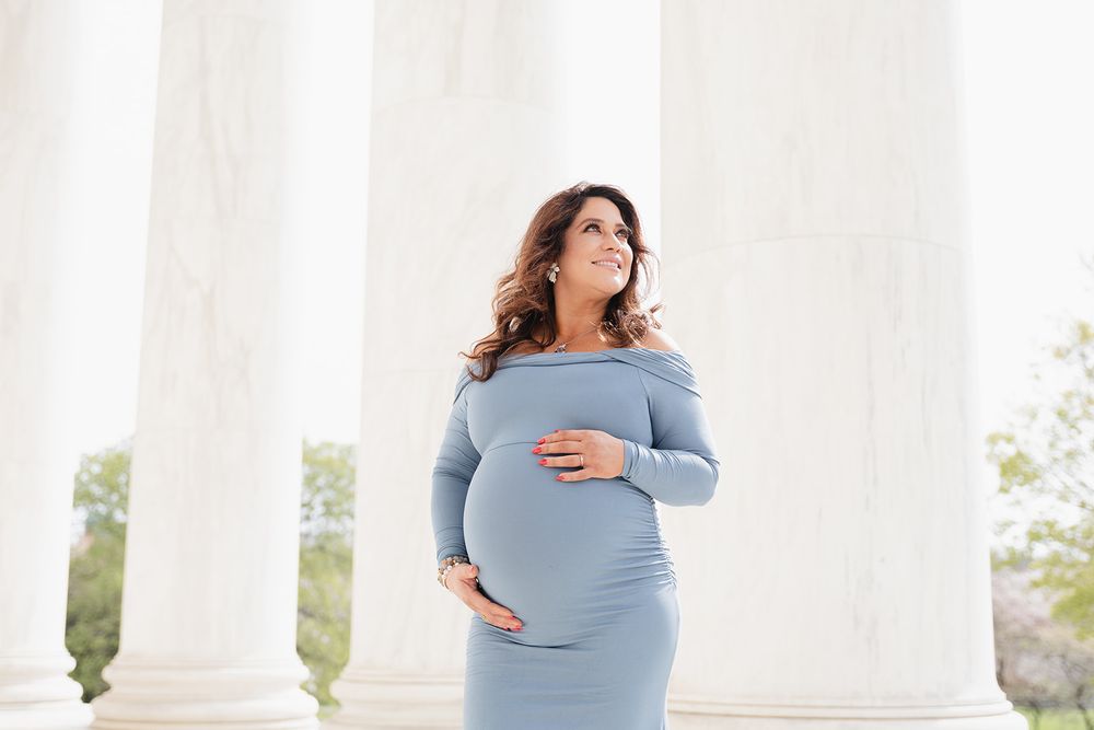 Maternity photo of an expectant mother in a light blue off-shoulder dress posing elegantly between white columns.