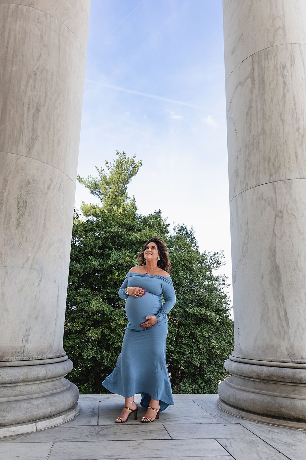 A person in a blue dress poses creatively between white marble columns outdoors against a blue sky.