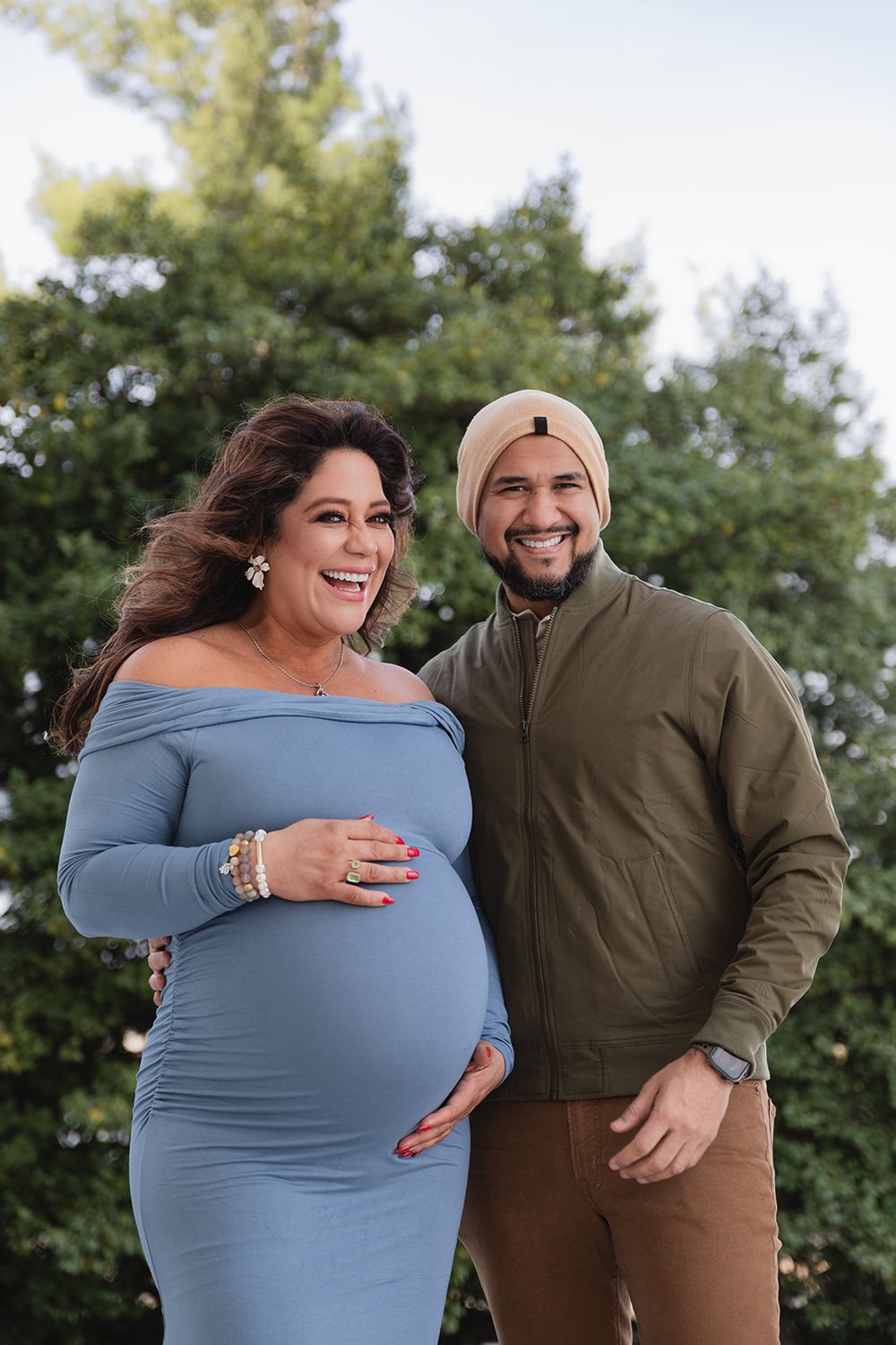 A couple shares a joyful moment outdoors, laughing and embracing while wearing casual clothing against a green backdrop.