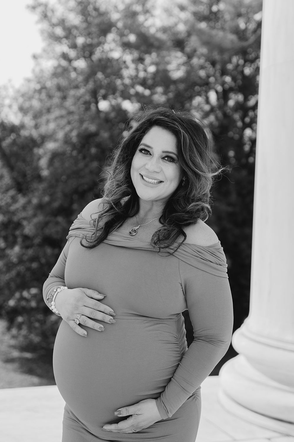 Black and white maternity portrait of an expectant mother in an off-shoulder dress smiling outdoors with natural lighting.