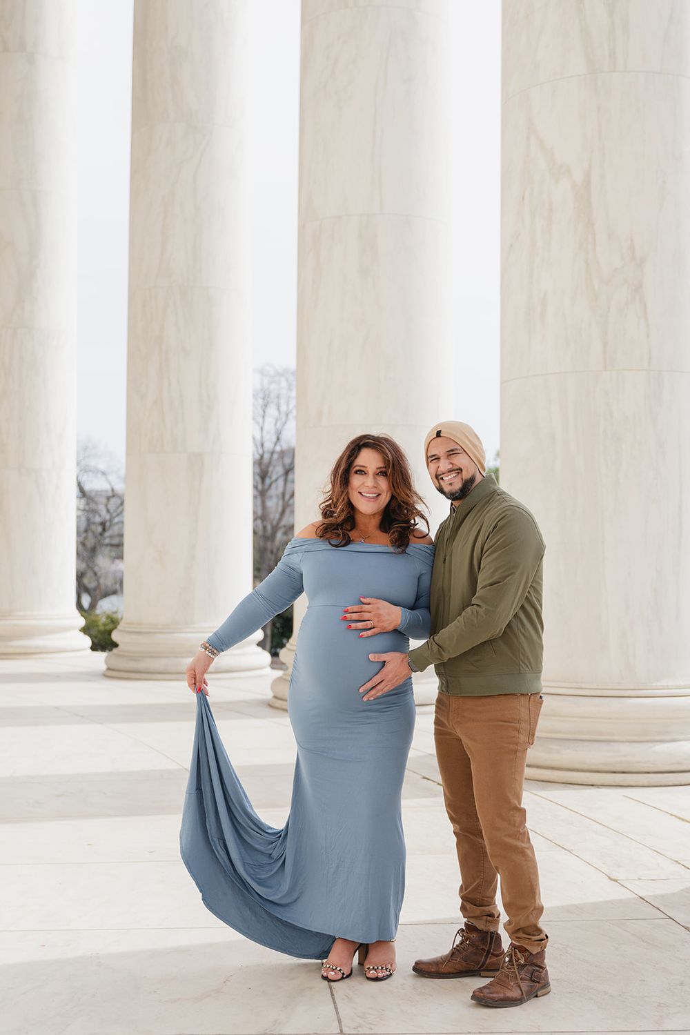 A couple poses playfully at a columned monument during a maternity photoshoot in soft blue attire.