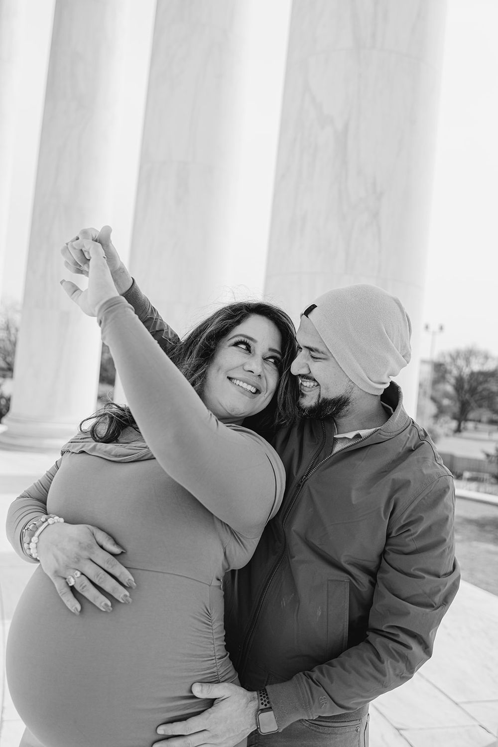 A joyful expecting couple embraces in front of white columns during a black and white maternity photoshoot.