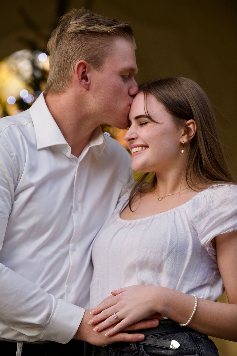 Man kissing smiling woman on forehead showing engagement ring outdoors.