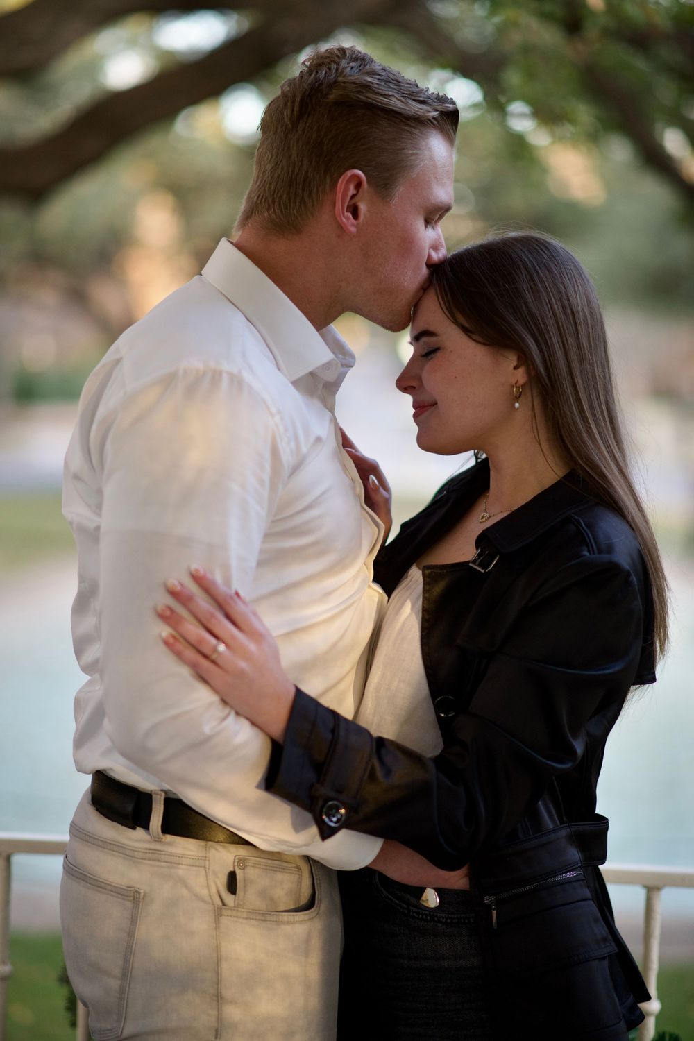 A couple shares a tender forehead kiss while embracing outdoors in soft evening light.