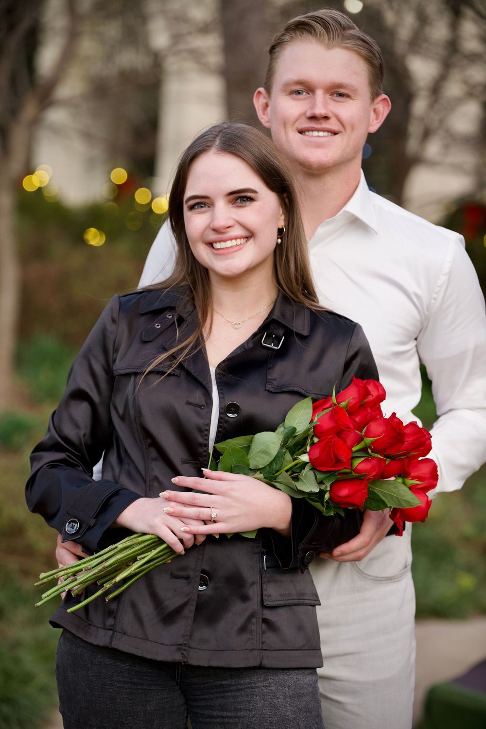 A smiling couple poses outdoors, the woman holding a bouquet of red roses with an engagement ring visible.
