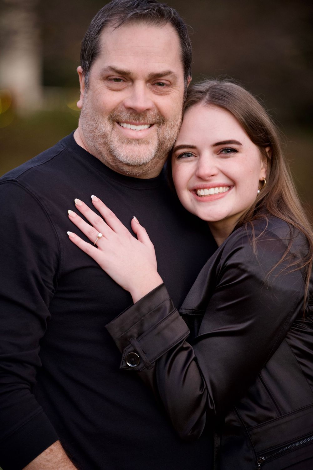 Smiling man and woman in black outfits embracing outdoors, woman showing engagement ring.
