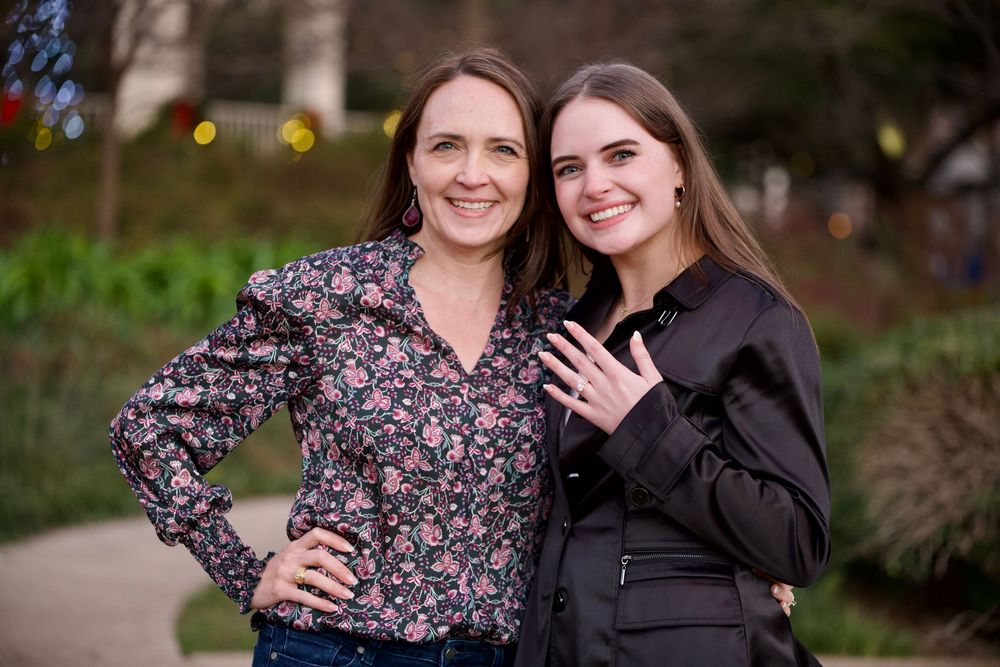 Two smiling women posing together outdoors, one displaying an engagement ring.