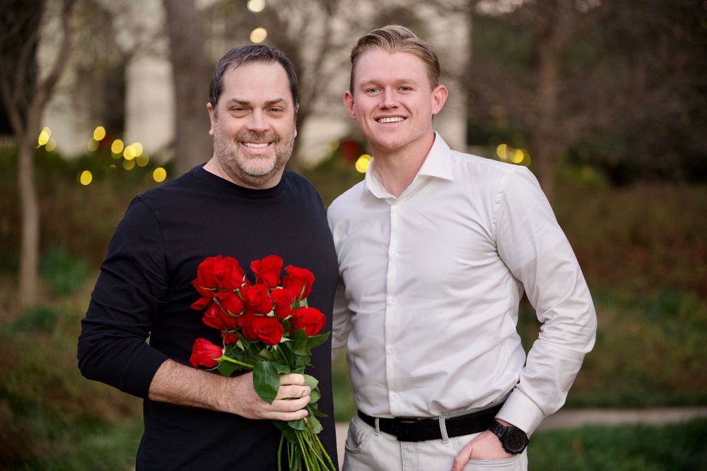 Two smiling men posing outdoors, one holding a bouquet of red roses.