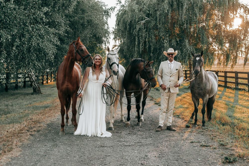 Ellensburg, WA wedding couple posing with their horses on a gravel road with trees and fences lining the sides