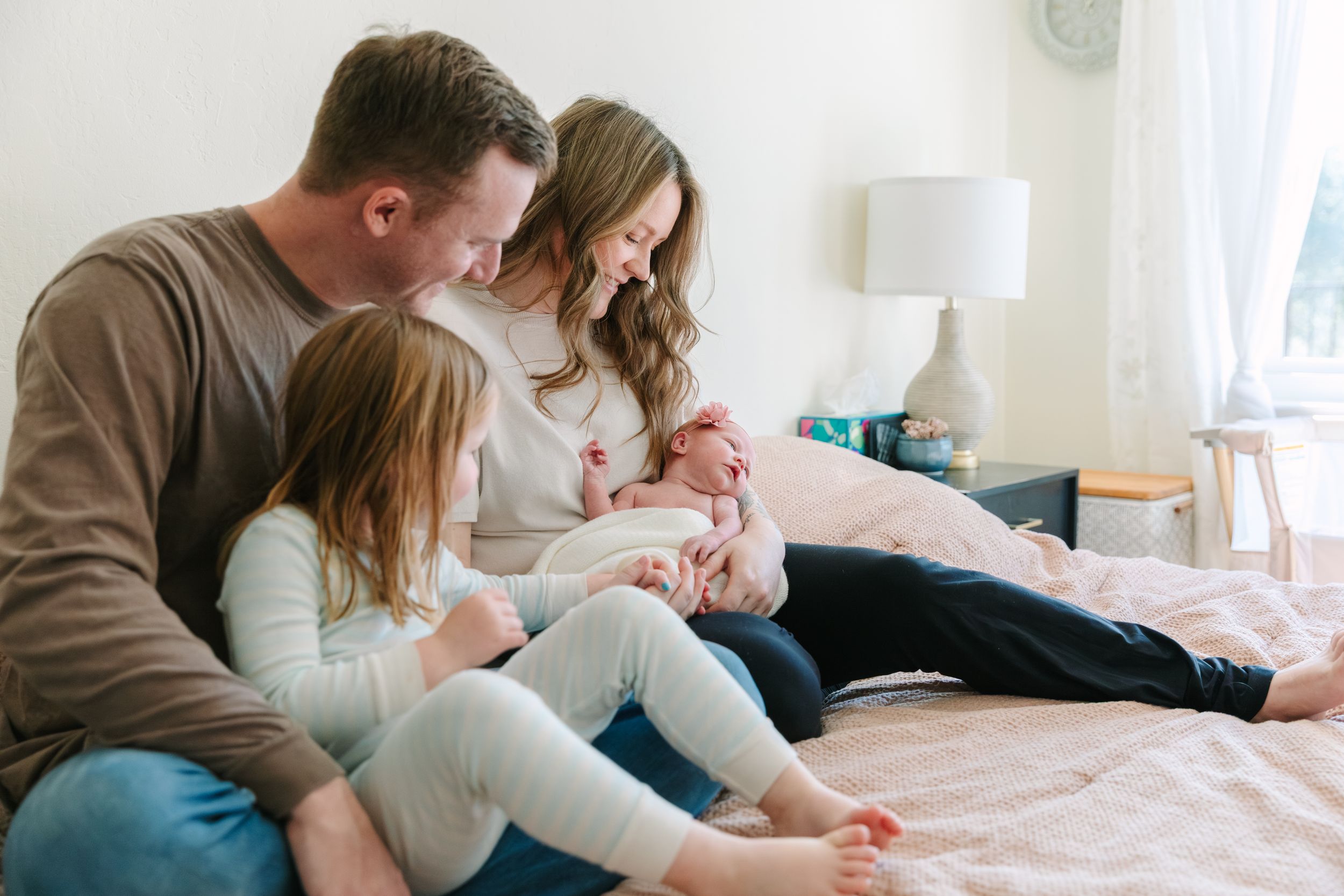 A family lounges together on a bed while bonding with their newborn baby in a bright, airy bedroom.