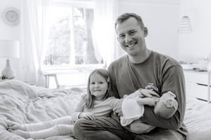 Family relaxing together on white bedding in a bright sunlit bedroom.