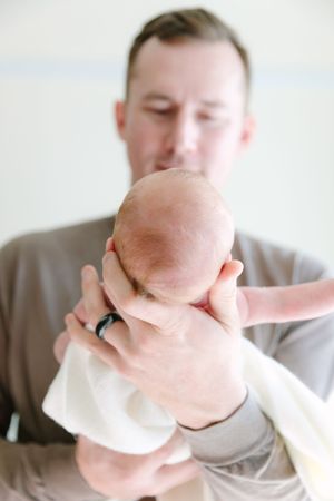 A person in a brown sweater gently holds and gazes at a newborn baby wrapped in white.