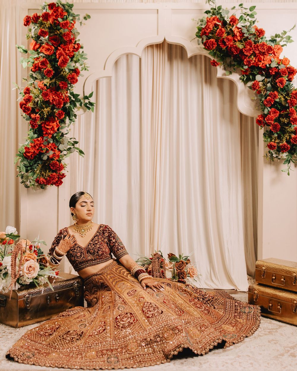 Ornate bridal lehenga with intricate embroidery photographed against white curtains and red flowers.