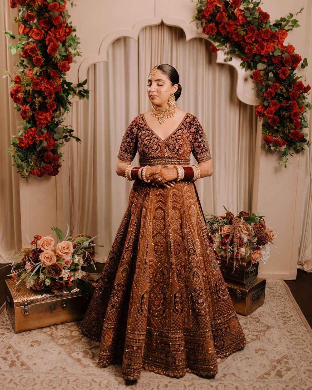 Intricate brown and gold embroidered wedding lehenga with red floral decor in elegant indoor setting.