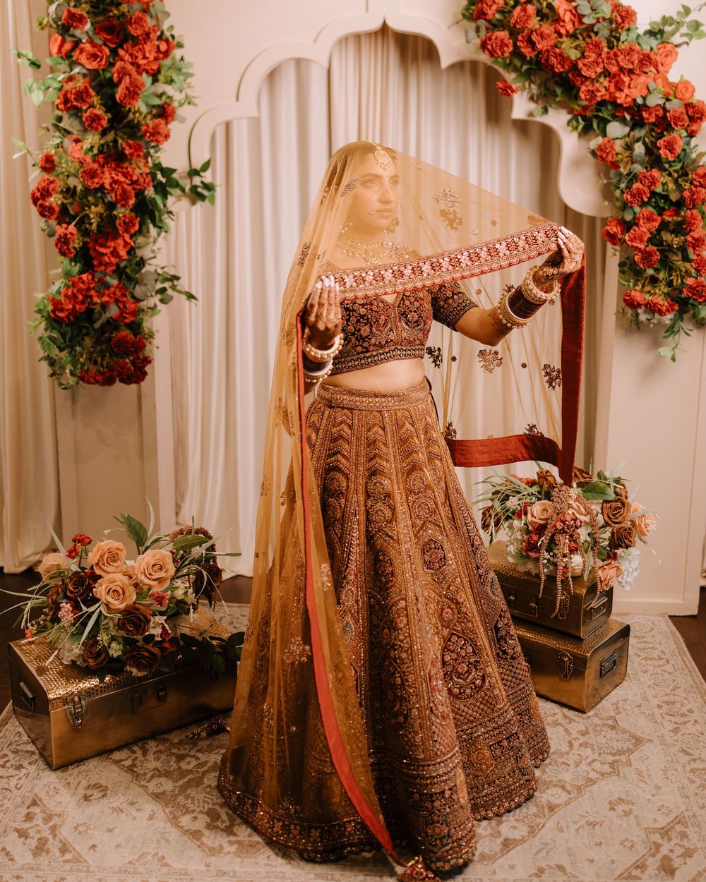 Ornate burgundy and gold Indian bridal lehenga displayed with red floral decorations and white drapes.