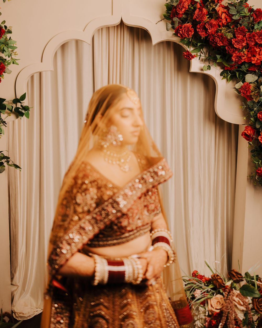 Traditional South Asian bridal attire with red floral decor and white curtain backdrop.