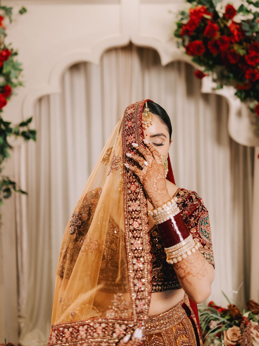 Traditional Indian bridal attire with gold embroidered lehenga and sheer veil against floral backdrop.