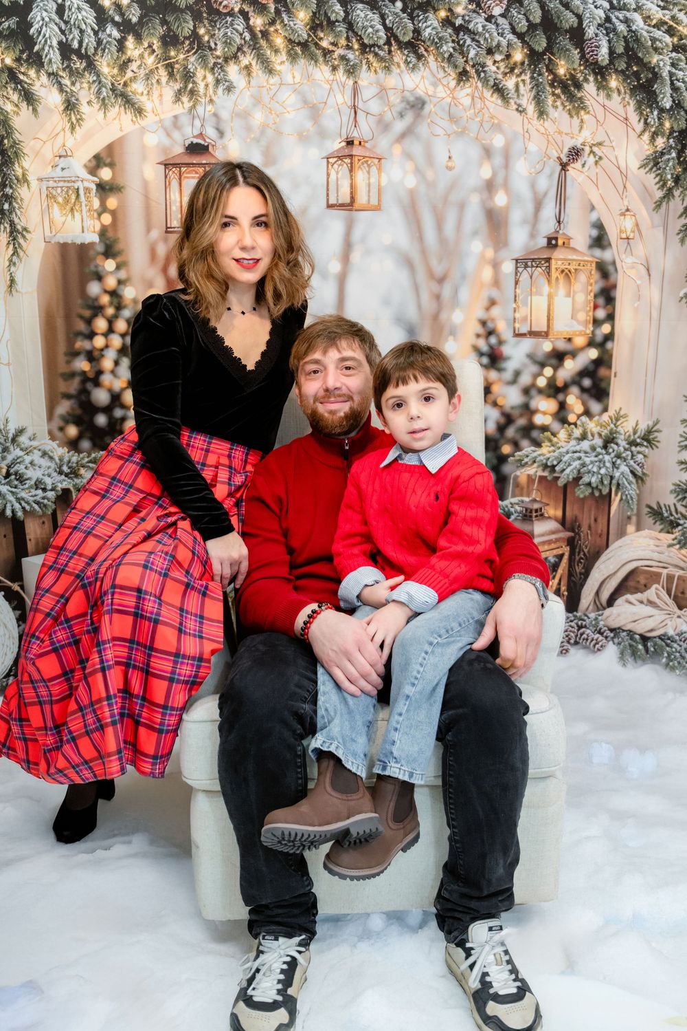 Family wearing coordinated red and black outfits pose together in winter wonderland setting with lanterns and snow.