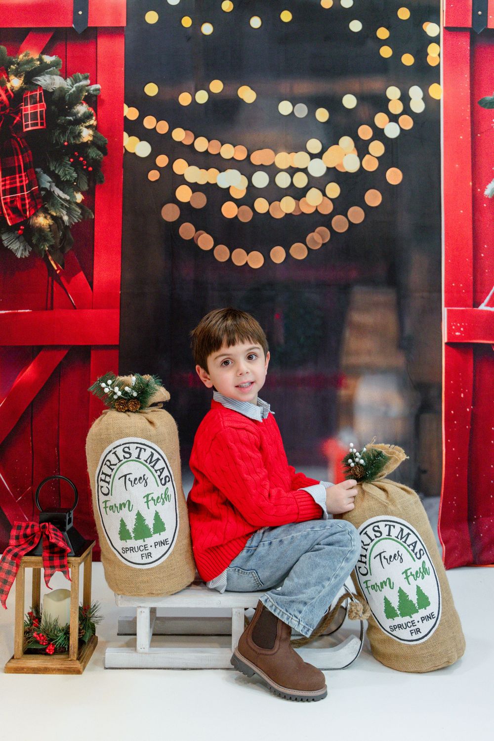 A festive holiday scene with a red window frame, decorative lights, and rustic tree farm signs displayed on a wooden bench.